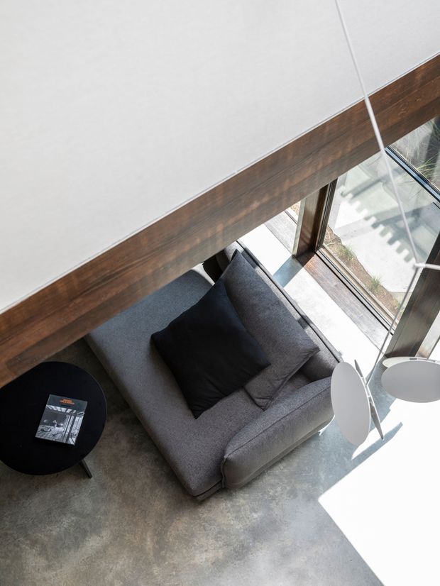 Overhead view of a compact contemporary living area with a grey upholstered sofa, black cushions, round black side table, and small white nesting tables. The space has a polished concrete floor, dark timber window framing, expansive glazing, and strong natural sunlight. A large white ceiling plane and timber beam are visible above the seating area.