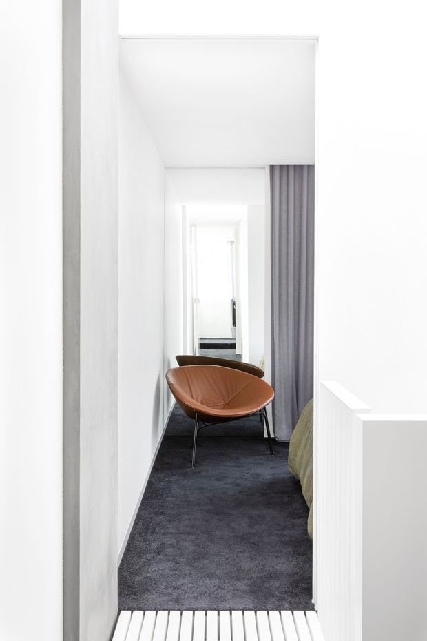 A narrow, bright white hallway or landing with dark charcoal carpet, a brown leather lounge chair, and a grey floor-length curtain beside an opening. White walls and ceiling create a minimalist, high-contrast interior, with repeated doorways or openings extending into the distance. A partial bed or upholstered furniture edge is visible at right, suggesting a bedroom adjacent to the passage.