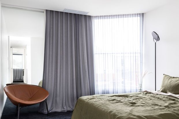 A bright, contemporary bedroom with a low-profile bed dressed in muted olive-green bedding, a sculptural brown leather lounge chair, full-height grey curtains, sheer floor-to-ceiling window coverings, and a slim black floor lamp. A mirrored sliding wardrobe or wall panel reflects the room, while dark carpet and restrained furnishings create a calm, minimalist atmosphere.