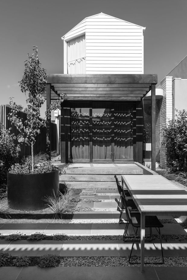 Black-and-white photograph of a compact contemporary townhouse facade and landscaped front courtyard. A dark timber pergola shelters glazed entry doors, with a stepped paved path, raised planter, ornamental planting, and an outdoor dining table in the foreground. The upper level is clad in horizontal white weatherboards with a simple pitched roof.
