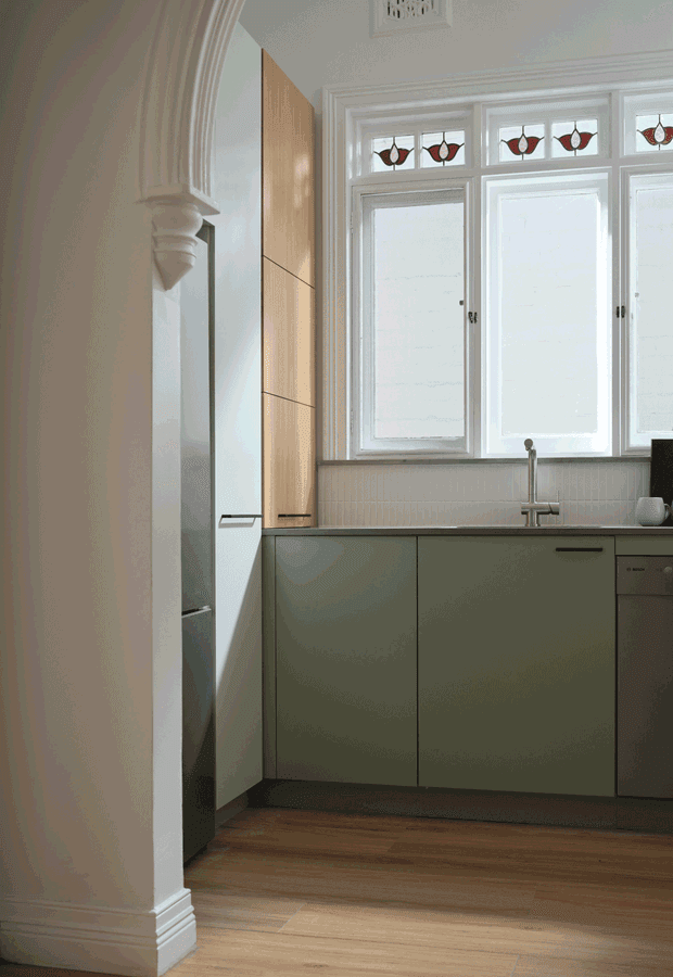A compact, refined kitchen viewed through an ornate arched opening. The room features muted green flat-front cabinetry, a timber-toned tall pantry or appliance cabinet, white benchtops and backsplash, integrated appliances, a stainless-steel tap, warm timber flooring, and tall white-framed windows with decorative stained-glass fanlights. Traditional architectural mouldings contrast with contemporary minimalist joinery.