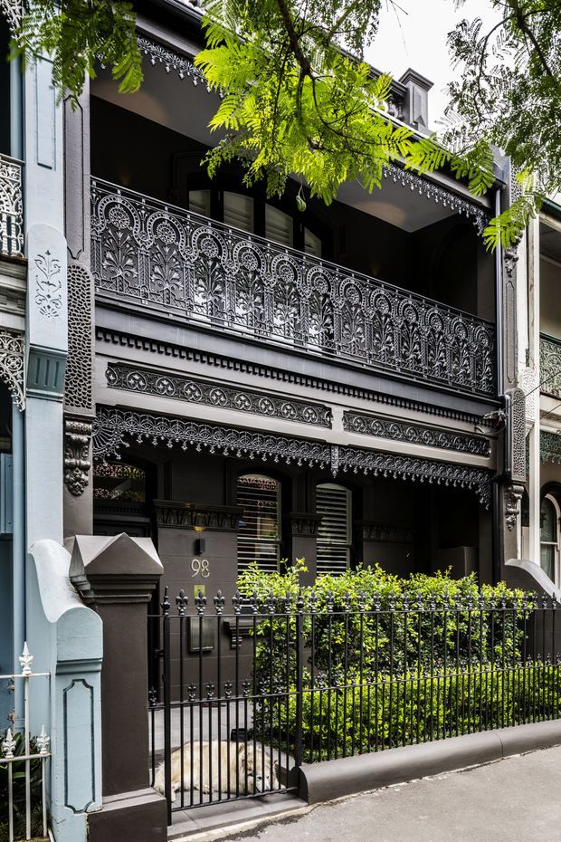 Street-level view of a narrow, multi-storey terrace house facade with highly ornate cast-iron lacework balconies, decorative brackets, patterned friezes, classical detailing, a dark painted exterior, and a small fenced front garden. Mature foliage partially frames the upper facade.