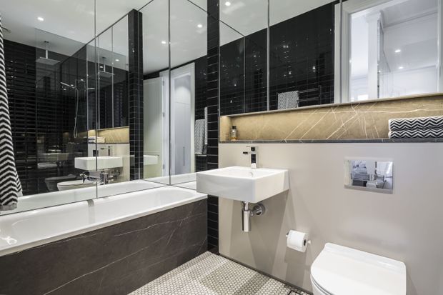 Contemporary bathroom with a freestanding bathtub clad in dark stone-look panels, wall-mounted white basin, concealed-cistern toilet, large mirrored wall, glossy black tiled feature walls, and a warm beige stone-look horizontal ledge beneath the window. Small light mosaic floor tiles and bright recessed lighting create a crisp, high-contrast interior.