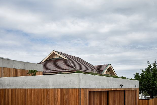 Exterior view of a suburban residential property, dominated by a tall timber-clad boundary wall and gate in the foreground. Behind it is a pitched, gabled house roof with reddish-brown tiled roofing and light-coloured gable detailing. The image has an overcast sky and a restrained natural material palette.