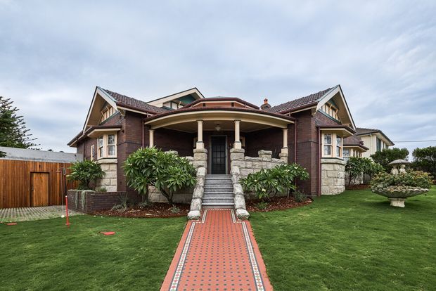 Front exterior view of a substantial period-inspired brick residence with a symmetrical entrance porch, stone-clad columns and steps, gabled roof forms, dormer-like upper windows, and a patterned red tile walkway leading through a manicured lawn. The home is presented in a traditional, formal style with mature garden planting and timber fencing.