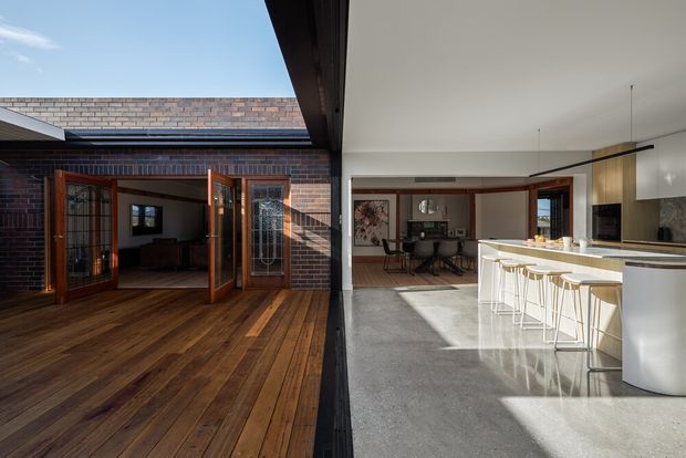 A contemporary open-plan kitchen and dining area opens onto a timber-clad courtyard deck through large glazed timber doors. The kitchen features a long white island with integrated seating, pale cabinetry and dark benchtops, while the adjacent dining area has a dark table and chairs. Exposed dark brick walls, timber decking, polished concrete flooring, high ceilings and strong indoor–outdoor connection define the space.