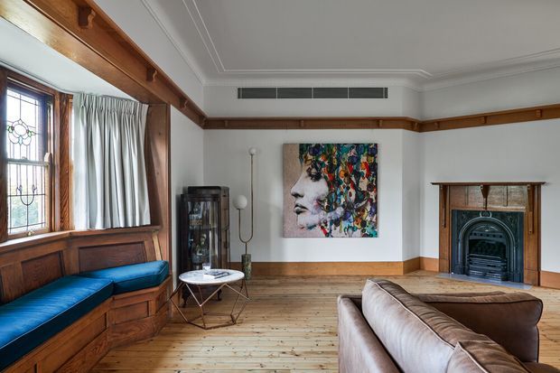 A refined traditional living room with extensive dark timber detailing, built-in window seating upholstered in deep blue, a timber floor, decorative ceiling cornices, and a classic fireplace with a dark metal insert. A contemporary colourful portrait provides a bold focal point alongside a leather sofa, glass display cabinet, and sculptural side table.