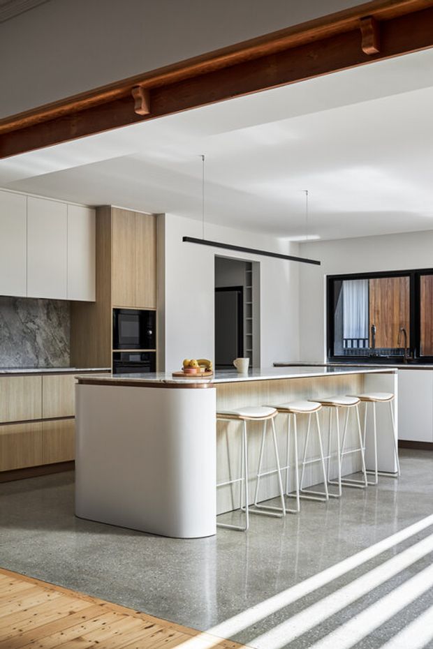 A bright contemporary kitchen with a large white waterfall island and integrated seating for four. The kitchen features light timber cabinetry, a dark natural-stone backsplash and benchtop accents, built-in appliances, polished concrete flooring, and a long slim black pendant light. Exposed dark timber ceiling beams and a timber-framed window add warmth and architectural character.