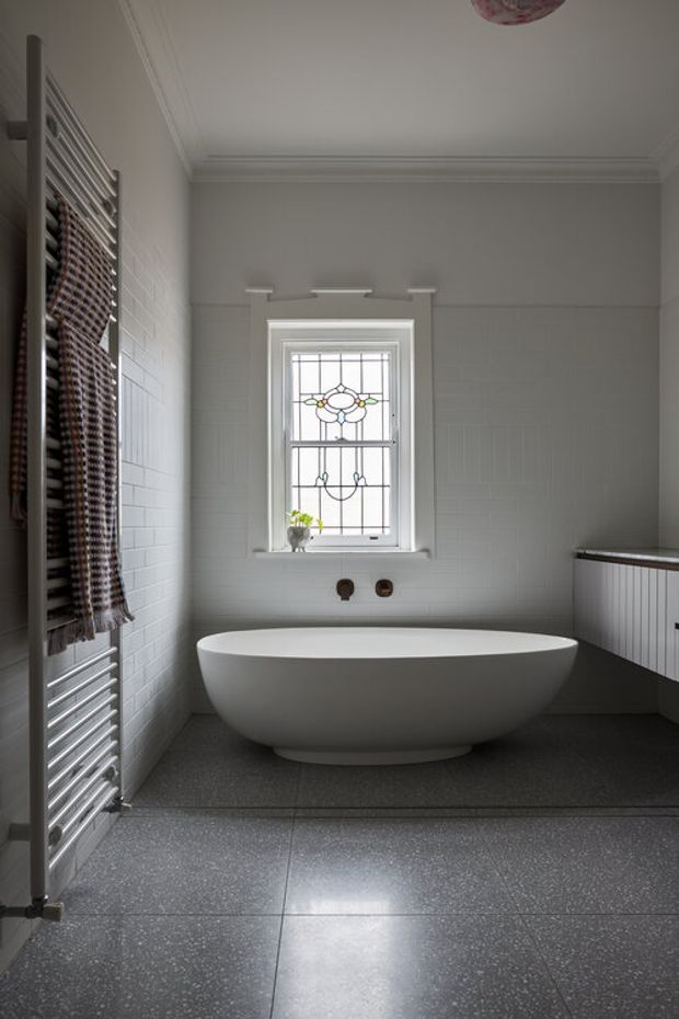 A refined bathroom featuring a freestanding white oval bathtub beneath a decorative leaded-glass window. The room has white tiled walls with traditional panel detailing, a dark grey tiled floor, a heated towel rail with a patterned towel, and white built-in cabinetry with a dark countertop. Ornate ceiling cornices and restrained period detailing create a calm, classic atmosphere.