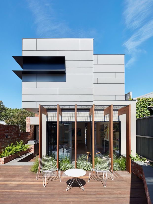 A narrow multi-storey contemporary residence viewed from the rear, with a crisp white panelled upper volume projecting over a glazed ground-floor living area. Vertical timber fins shade the glass facade, opening onto a spacious timber deck furnished with two wire lounge chairs and a small round table. Brick boundary walls, dark fencing, planted garden beds and a bright blue sky frame the house.