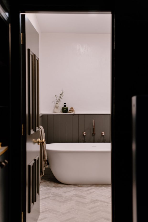 A refined bathroom viewed through a partially open dark-framed door. The room features a freestanding white bathtub, dark vertical-panel wall wainscoting with exposed copper-toned tapware, pale herringbone floor tiles, and a softly lit white upper wall. A small vase, soap dispenser, and folded towels add understated styling.