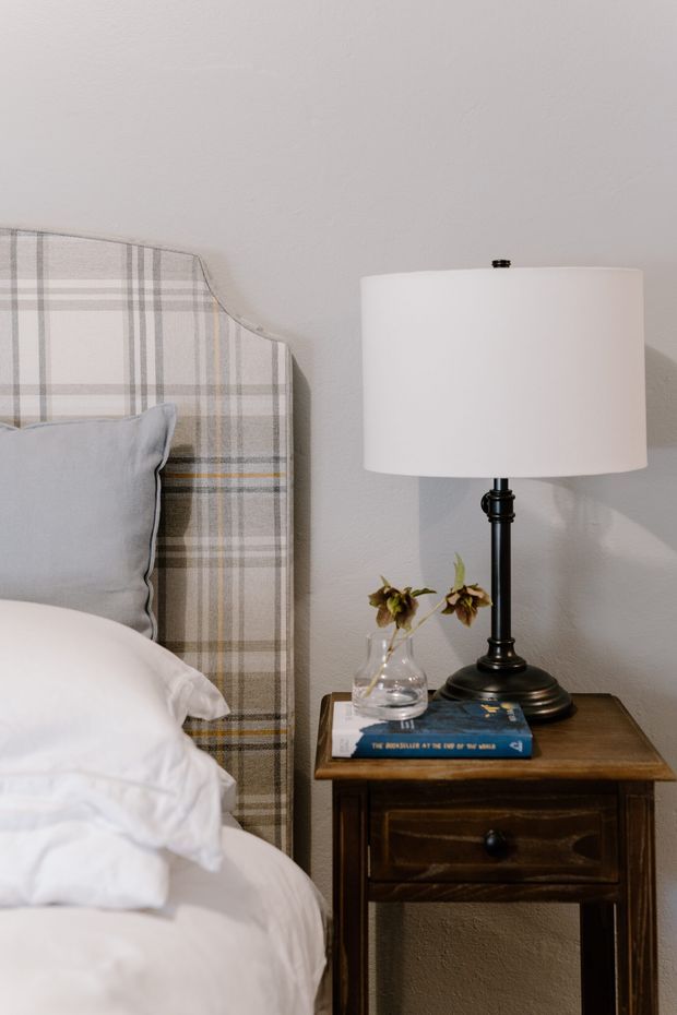 Close-up view of a traditional bedroom corner featuring a tall upholstered plaid headboard, white bedding, a pale blue-grey pillow, and a dark wood bedside table with a classic black table lamp, glass vase, and books. The room has a soft neutral palette and warm, intimate styling.