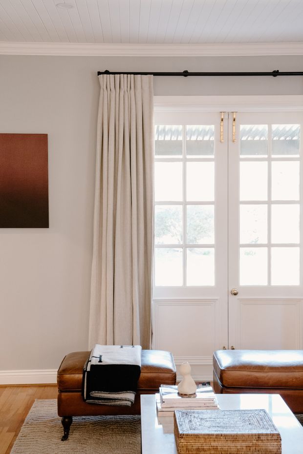 Bright, refined living room with white-painted French doors, full-height cream curtains on a black rod, a white beadboard ceiling, pale neutral walls, and timber flooring. Two cognac leather ottomans or benches sit around a glass coffee table with books and a woven basket, while a simple dark-toned artwork hangs on the wall.
