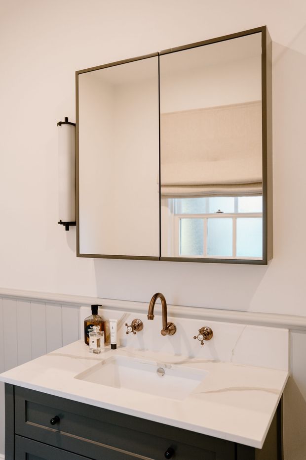 A refined bathroom vanity with a dark painted timber cabinet, white stone benchtop and integrated rectangular basin. Brass wall-mounted tapware and a large framed mirrored cabinet sit above the vanity, with light grey-blue vertical wall panelling and warm off-white walls creating a classic, softly lit interior.