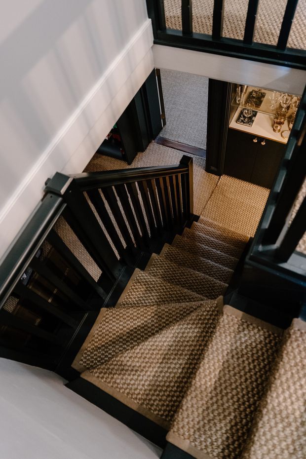 A top-down view of a stairway with dark painted timber balustrades and handrails, beige patterned carpet covering the stairs and landing, white walls with simple trim, and a black built-in cabinet displaying decorative objects at the lower level.