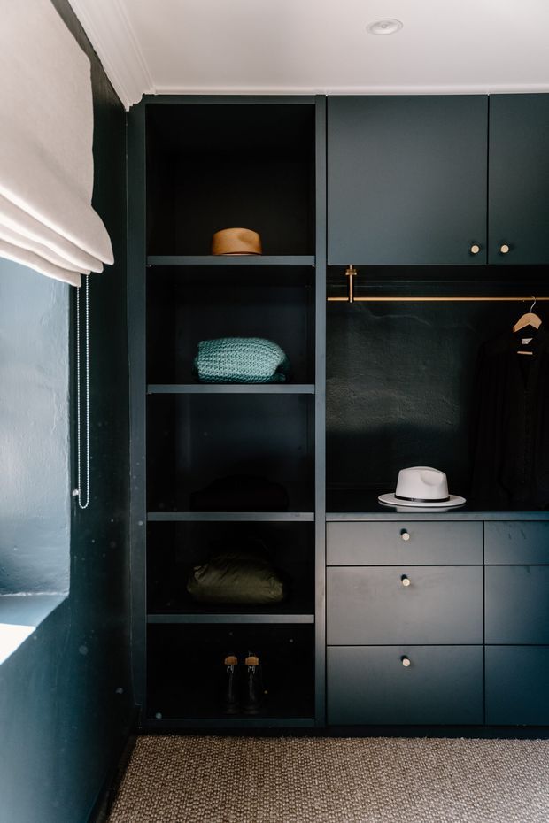 A dark, richly coloured built-in wardrobe and dressing storage wall fills the image. Matte deep blue-green cabinetry includes open shelving, drawers, a hanging rail with clothing, and integrated display areas. A white Roman blind sits beside a window on the left, with a textured neutral carpet underfoot and crisp white cornice detailing above. Brass-toned hanging hardware and small round knobs add subtle contrast.