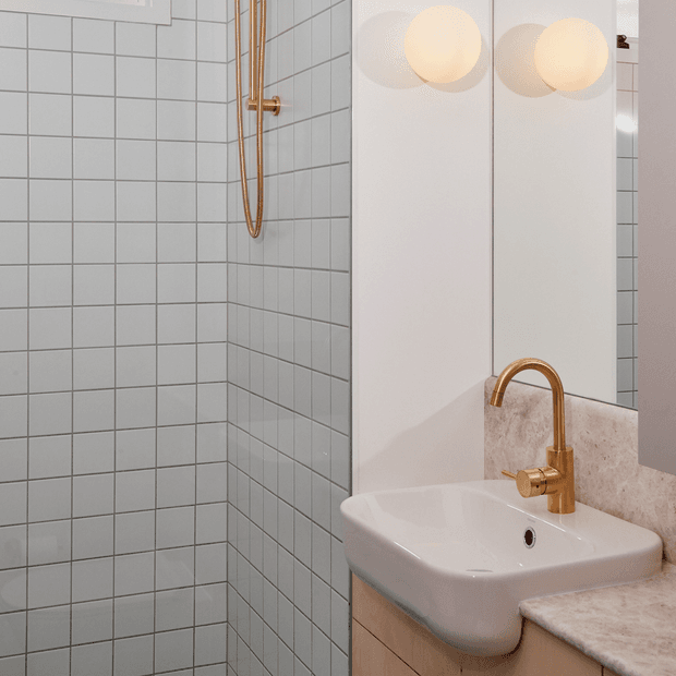Compact contemporary bathroom featuring a wall-mounted white basin on pale wood-toned cabinetry, a beige stone-look benchtop and splashback, square light-grey wall tiles, brushed brass tapware and shower fittings, and a large mirror with globe wall lights.