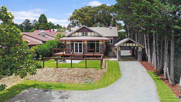A standalone suburban house viewed from the driveway, featuring a gabled roof, cream weatherboard-style exterior with dark trim, a covered front veranda, and a detached carport or garage structure. A gravel driveway curves through a landscaped front garden, with mature trees and neighbouring homes surrounding the property.