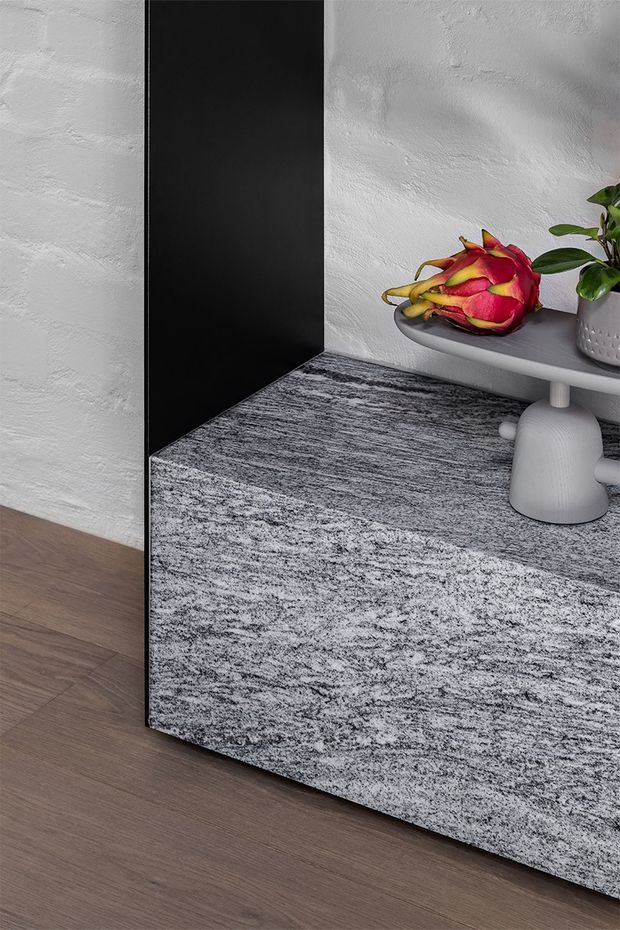 Close-up interior detail showing a large freestanding or built-in grey-and-black textured rectangular cabinet or plinth against a white textured wall. A grey side table with a potted plant and dragon fruit sits behind it. The surrounding floor is medium brown timber, while the cabinet face has a mottled stone-like or textured finish.