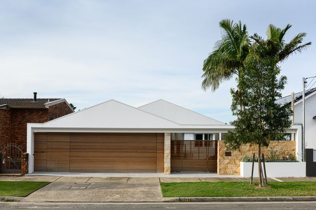Street-facing view of a contemporary single-storey residence with a broad low-pitched gable roof, white rendered facade, warm horizontal timber garage door and matching timber screening, stone-clad entry wall, covered porch, and manicured front lawn. Mature palm trees and neighbouring homes frame the suburban setting.