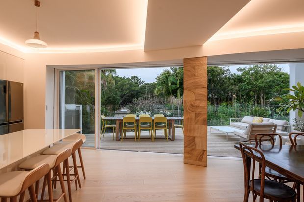 Open-plan contemporary kitchen, dining and living area with extensive sliding glass doors opening to a landscaped terrace. The kitchen has a large pale island with timber bar stools, integrated dark appliances and light cabinetry. A timber-clad structural column separates the dining area from the lounge, while recessed cove lighting creates a warm, softly illuminated interior. Outdoor dining and greenery are visible beyond the glazing.