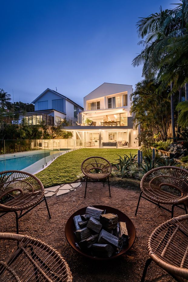 A dusk-time view across a landscaped backyard with a swimming pool, lawn, tropical planting, and a circular outdoor fire pit surrounded by woven lounge chairs. A large contemporary multi-level house with extensive glazed living areas and illuminated balconies forms the main architectural backdrop, with a neighbouring gabled home visible to the left.