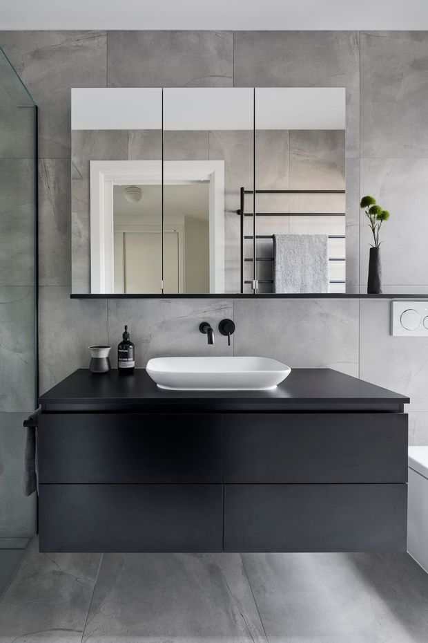 A contemporary bathroom vanity photographed straight on. Large grey concrete-look tiles cover the walls and floor, with a floating matte-black vanity topped by a black benchtop and a white vessel basin. Above it is a wide frameless mirrored cabinet, while a black ladder-style heated towel rail, folded white towel, black tapware, wall-hung toilet flush plate and partial bathtub are visible. The palette is restrained in grey, black and white with clean, minimal lines.