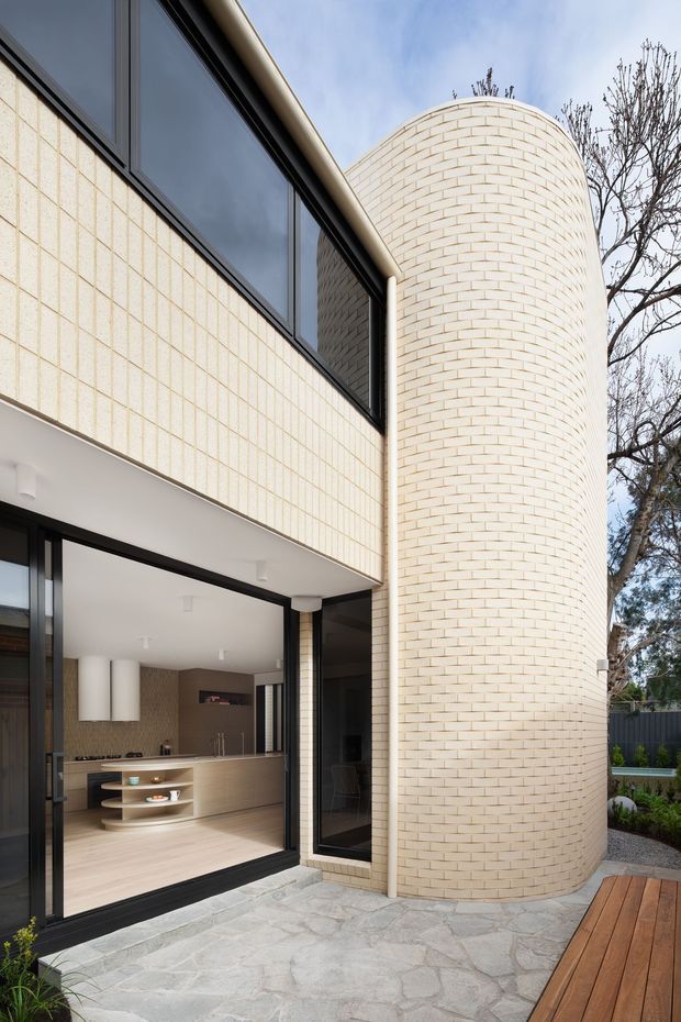 Exterior view of a contemporary two-storey residence with pale cream brickwork, a prominent curved brick tower, large black-framed glazing, and an open-plan kitchen visible through the ground-floor doors. The building opens onto a paved courtyard with a timber deck and landscaped garden.
