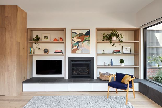 Contemporary living room featuring a built-in timber media wall with open shelving, a central black fireplace, low white storage cabinetry, and a navy upholstered armchair. Large corner glazing brings in natural light, while curated books, plants, ceramics, and artwork add warmth.