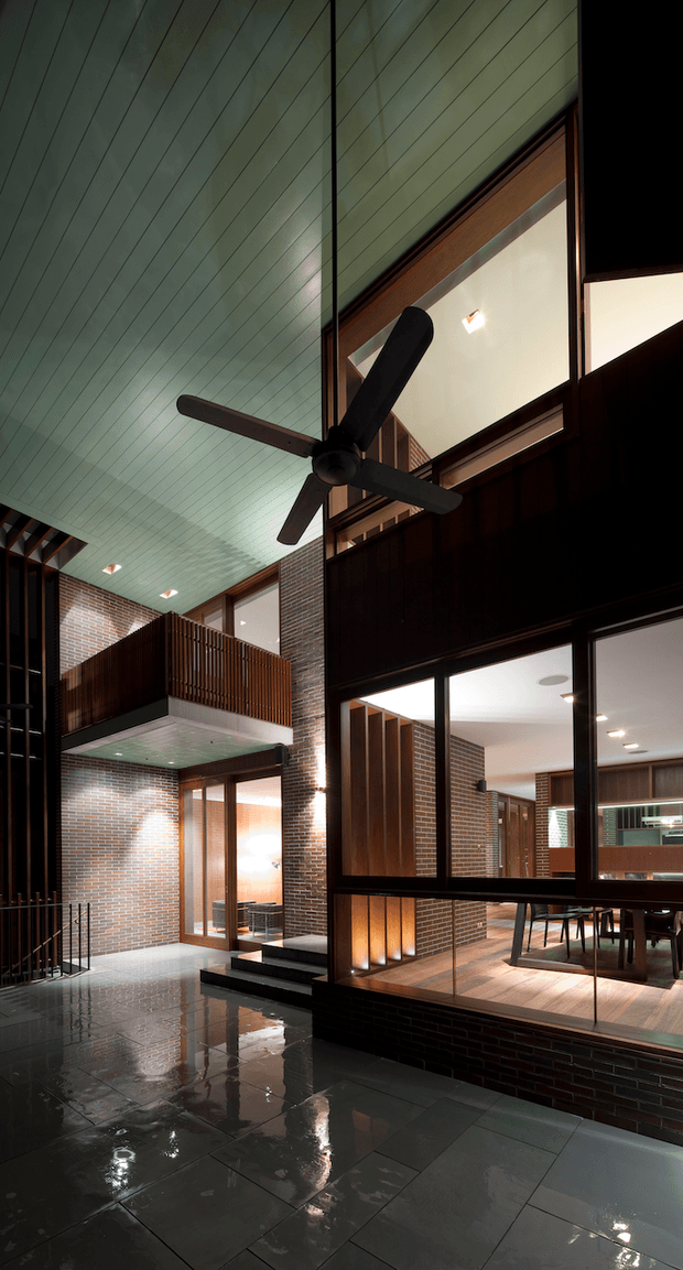 A dramatic interior courtyard or atrium of a multi-level modern residence, viewed from a polished dark tiled floor. The space features exposed red brick walls, dark timber screens and balustrades, extensive black-framed glazing, a pale timber-lined ceiling, and a large black ceiling fan. A dining area and warm-lit interior rooms are visible through the glazing, while a balcony and stair access add vertical layering.
