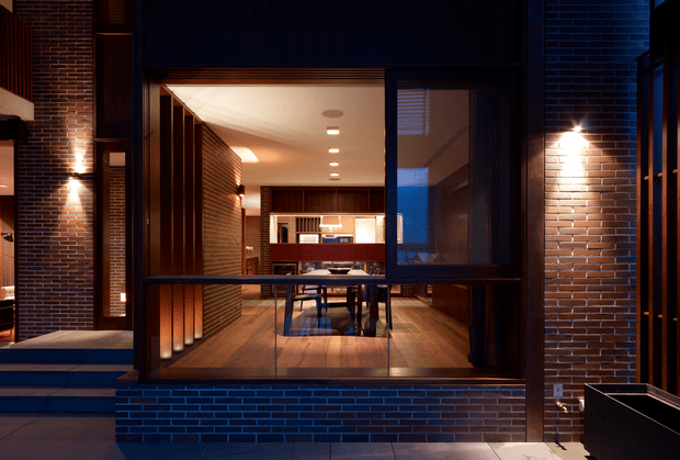 A twilight exterior view through a large glazed opening into a warm, contemporary brick residence. The dark brick facade is accented by wall-mounted lighting and black-framed glazing. Inside, a timber-floored dining area with a table and chairs leads toward a kitchen or built-in joinery, with timber slatted wall elements and recessed ceiling lights creating a warm, intimate atmosphere.