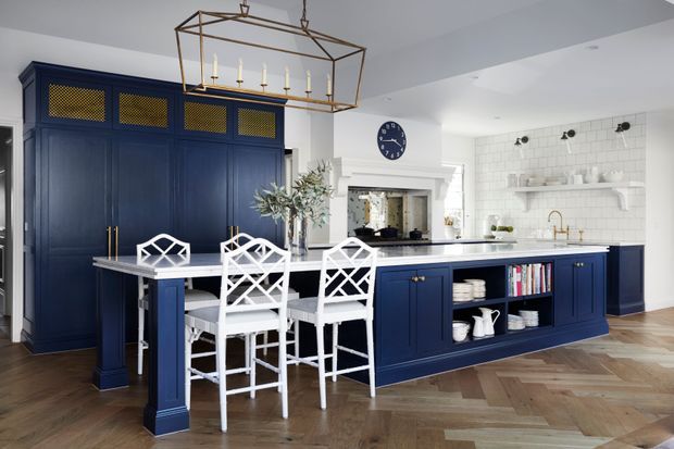 A spacious, bright transitional kitchen with deep navy-blue cabinetry, a large central island with white cross-back bar stools, white subway-tile backsplash, brass hardware and fixtures, open display shelving, and a geometric brass pendant light. The warm herringbone timber flooring contrasts with the crisp white walls and ceiling.