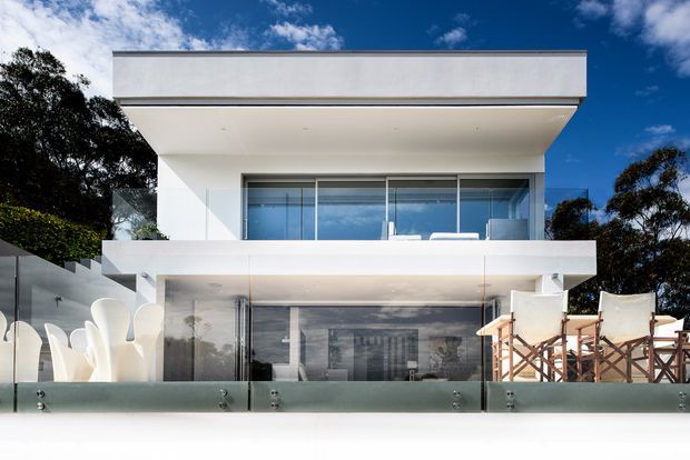 A modern multi-storey residential facade with crisp white rendered volumes, broad cantilevered slabs and extensive floor-to-ceiling glazing. A glass balustrade encloses the outdoor terrace, with sculptural white outdoor seating on the left and timber-framed deck chairs on the right. Mature trees and a blue sky form the background.