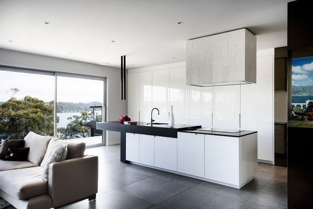 Bright open-plan contemporary kitchen and living area with a white island, glossy white cabinetry, black benchtops, integrated appliances, and a textured suspended rangehood enclosure. Floor-to-ceiling glazing opens views toward a waterfront landscape, while a neutral sofa occupies the foreground. The space has a clean, minimalist appearance with strong black-and-white contrast.