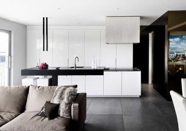A bright, open-plan contemporary interior combining a white minimalist kitchen with a black island and dark tiled flooring, opening into a living area with a taupe sofa. Glossy white cabinetry, a dark stone-look benchtop, slim black pendant lights, and a textured oversized feature pendant create a crisp, high-contrast scheme. A dark built-in wall with display shelving is visible to the right.