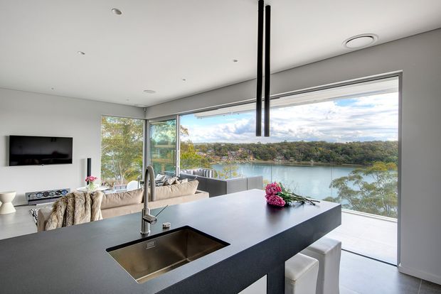 A bright, contemporary open-plan kitchen and living area with a large dark island featuring an integrated stainless-steel sink and white bar stools. Floor-to-ceiling glazing opens to a broad waterfront view, while a pale sofa, wall-mounted television, recessed ceiling lights, and slim black pendant lights complete the interior.