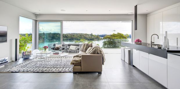 Bright open-plan contemporary living room and kitchen with expansive floor-to-ceiling glazing overlooking a waterfront landscape. The room features a large neutral sectional sofa, patterned rug, low coffee table, white minimalist kitchen cabinetry, dark island benchtop, integrated appliances, and polished large-format grey flooring. The space is airy, refined, and strongly connected to the view.