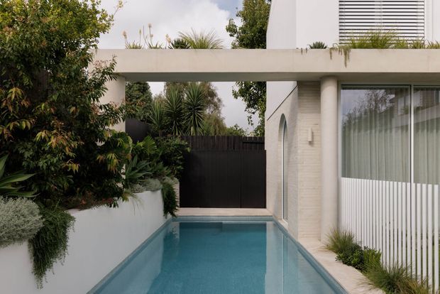 A narrow rectangular swimming pool runs alongside a modern white-rendered residence, framed by lush planting, a dark privacy gate, and a covered concrete walkway or canopy. Vertical glazing with sheer curtains and horizontal exterior blinds are visible on the right, while textured pale wall cladding and clean geometric forms create a restrained contemporary exterior.