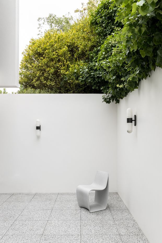 A minimalist enclosed courtyard with smooth white rendered walls, integrated black-and-white wall sconces, a sculptural white chair, and a speckled grey tiled floor. Dense green foliage spills over the upper walls, creating a strong contrast with the restrained architectural setting.