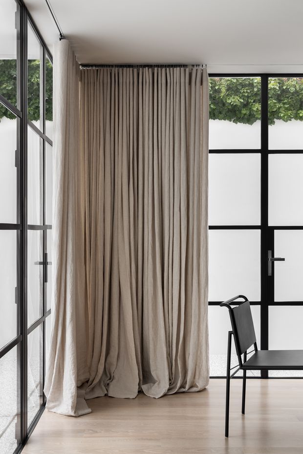 A bright, minimalist interior corner with full-height black-framed glazing and a matching black-framed door. Floor-to-ceiling taupe-beige curtains hang in soft folds from a ceiling track, pooling slightly on a pale timber floor. A simple black chair and table occupy the foreground, while leafy greenery is visible outside through the glazing.