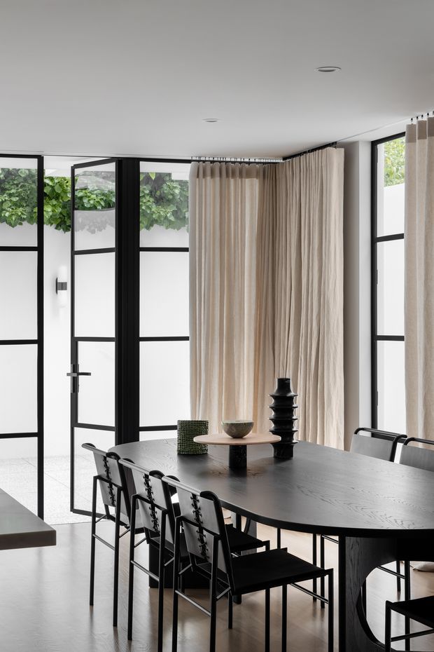 A refined dining room with a long black timber dining table and matching black-framed chairs. Full-height black-framed glazing and doors line the room, with sheer beige curtains gathered along the corner. White walls and ceiling, pale flooring, and restrained sculptural table decor create a monochromatic, elegant interior.