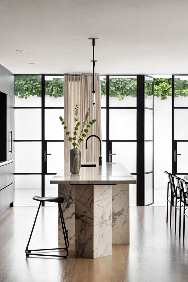 Bright contemporary kitchen centred on a sculptural marble island with an integrated sink and matte black gooseneck tap. The island has a waterfall-edged stone benchtop and matching marble-clad support, with a single black bar stool in the foreground and additional stools at the right. Black-framed glazed doors and windows line the rear wall, partially screened by full-height light curtains and greenery outside. Minimal white cabinetry, pale timber flooring, recessed ceiling lights and black pendant lighting create a clean, high-contrast interior.