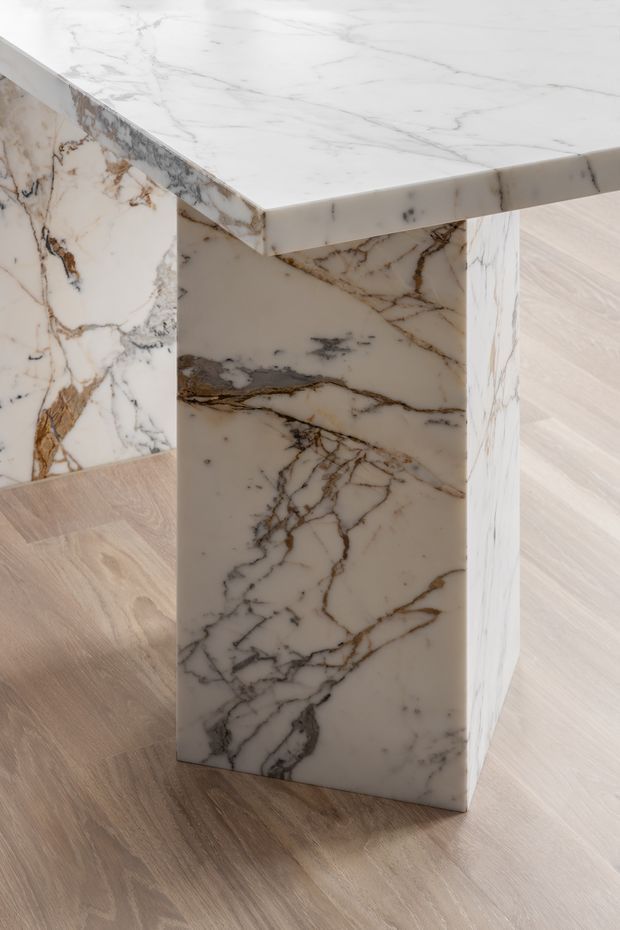 Close-up architectural detail of a substantial natural stone pedestal or island support with a matching thick stone top. The white stone has pronounced grey, charcoal, and warm brown veining, creating a luxurious sculptural feature. It stands on a pale timber-look floor in a softly lit interior.
