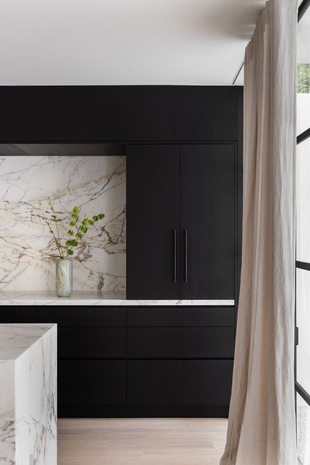 A refined contemporary kitchen with extensive matte black built-in cabinetry, long vertical handles, and a dramatic pale marble backsplash and benchtop with prominent veining. The edge of a matching marble island is visible in the foreground. Light timber flooring, full-height windows, and sheer beige curtains soften the dark joinery, while a small vase with greenery adds a natural accent.