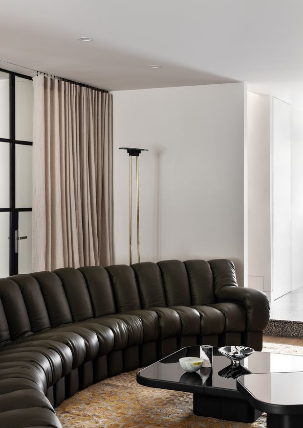 A contemporary living room with a large dark brown to near-black tufted sectional sofa, black-framed glazing, full-height beige curtains, a slim brass-and-black floor lamp, and sculptural black glossy coffee tables. A textured neutral patterned rug adds warmth, while white walls, a low white ceiling, and abundant daylight create a refined, high-contrast interior.