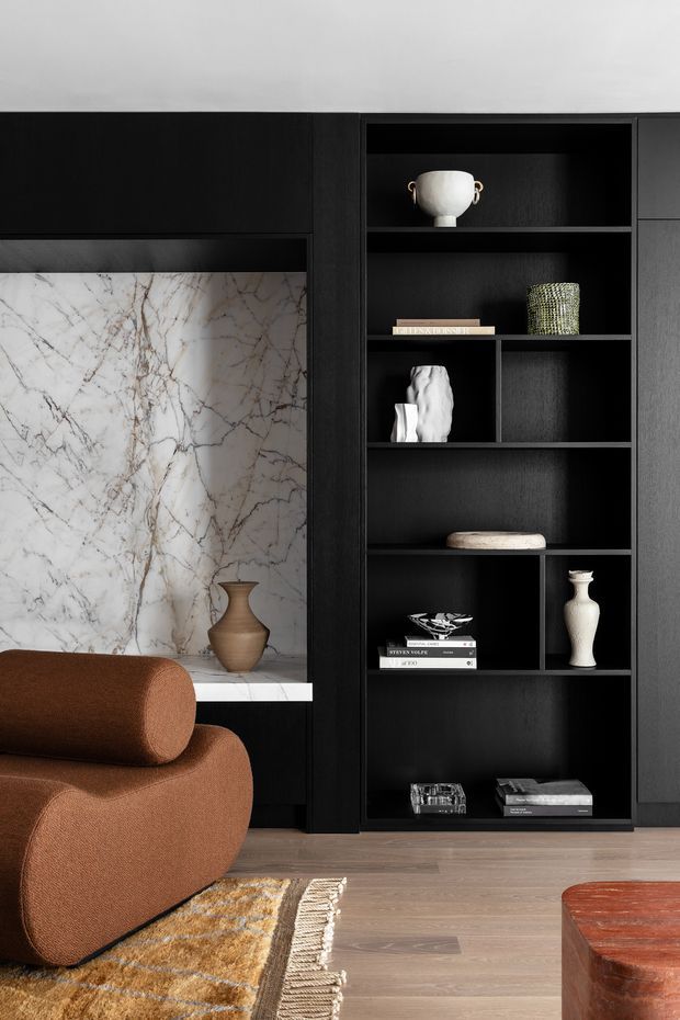 Close-up interior view of a refined living room featuring a floor-to-ceiling black built-in shelving and cabinetry wall, with open display niches styled with ceramics and books. A dramatic white veined marble feature panel sits beside the joinery. The foreground includes a curved rust-brown upholstered sofa, patterned rug, timber flooring, and a rounded wooden coffee table. The composition is highly curated with strong dark-and-light contrast and warm natural accents.