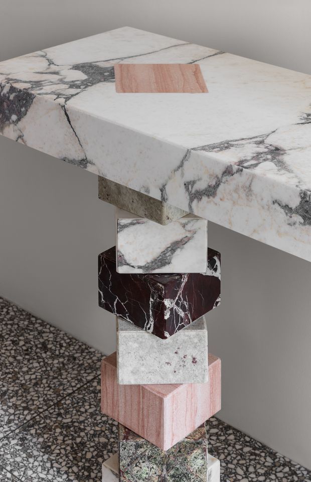 A sculptural display of stacked natural stone samples supporting a large veined stone slab, photographed against a neutral grey wall. The composition showcases contrasting marbled white, burgundy, pink, green and terrazzo-like stone surfaces, with a small pink sample placed on the upper slab. A terrazzo-style floor is visible below.