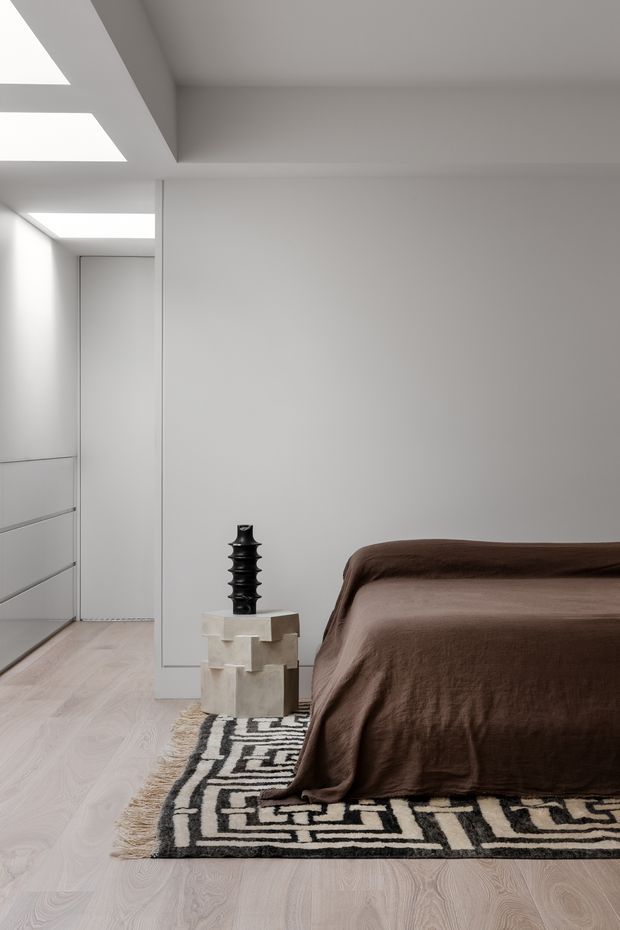 A minimalist bedroom with a low-profile bed dressed in dark brown bedding, set against smooth white walls and pale timber flooring. A black sculptural vase sits on a small light stone or concrete side table, with a black-and-cream geometric rug beneath the bed. Built-in white cabinetry and a concealed doorway line the left side, while a deep ceiling reveal and skylight-like openings bring in soft natural light.