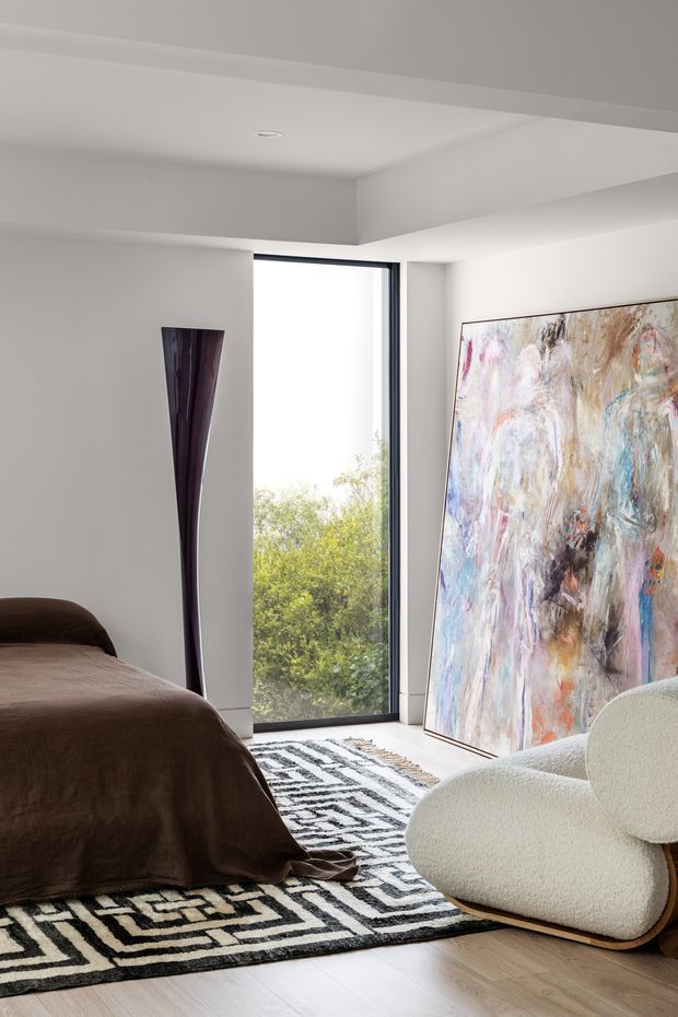 A contemporary bedroom with a low upholstered bed dressed in dark brown bedding, a sculptural dark floor vase, a large abstract artwork leaning against the wall, and a rounded cream lounge chair. A tall black-framed window provides abundant natural light and views of greenery. The room has white walls and a stepped ceiling detail, pale timber flooring, and a bold black-and-white geometric rug.