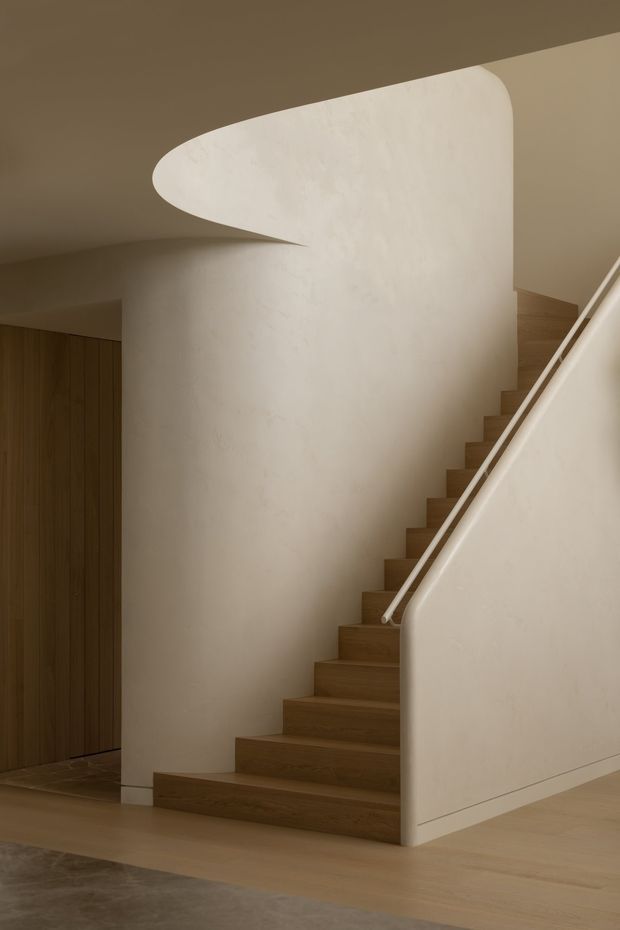 A refined interior stairway with warm timber treads rising beside a sculptural, curved plaster wall and integrated balustrade. The space has smooth off-white walls, a pale timber floor, dark timber cabinetry or wall panelling to the left, and soft, warm ambient lighting. The minimalist composition emphasizes the sweeping curved geometry and clean built-in detailing.